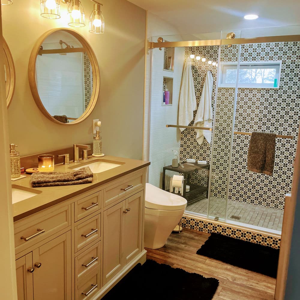 Modern bathroom with round mirror, glass shower, mosaic wall tiles, and wooden flooring.